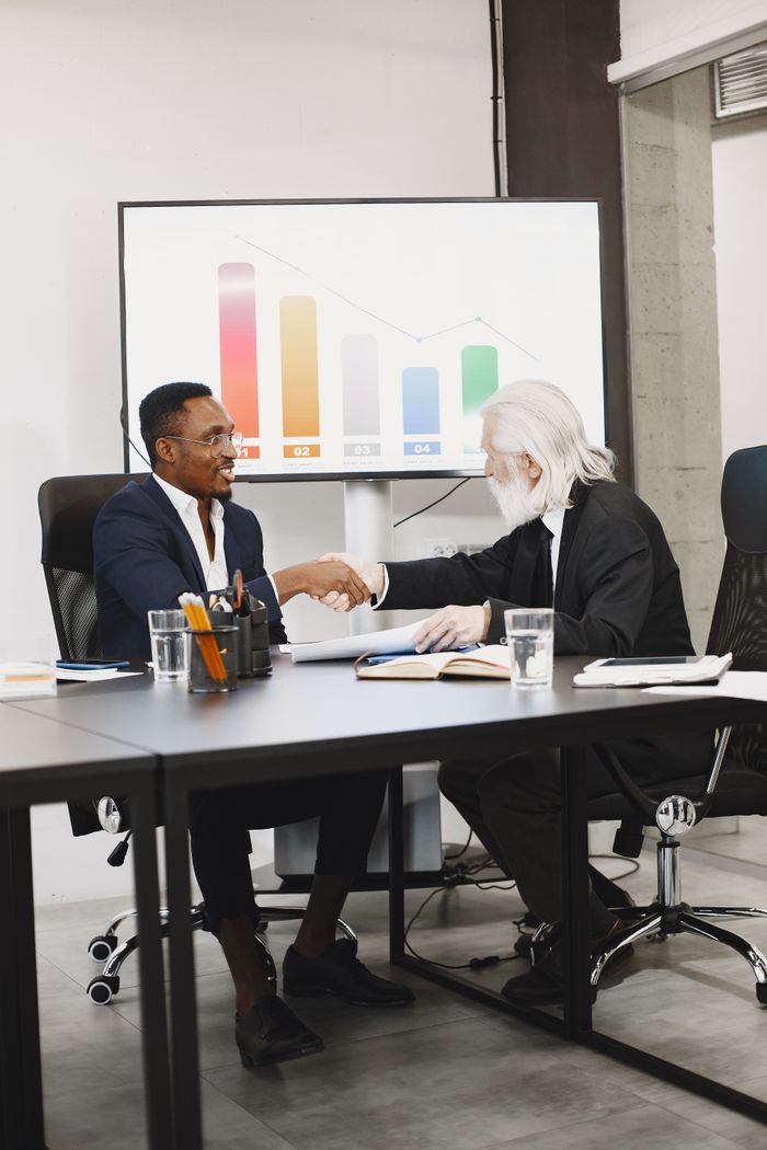 about-us-02 Two diverse businessmen in suits shaking hands over a desk in a modern office.